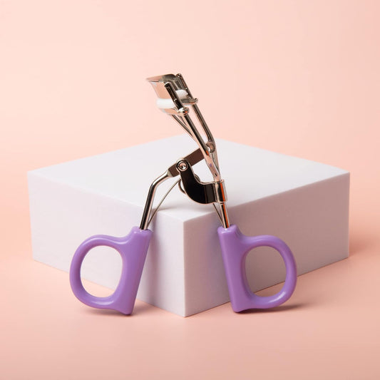 Professional Eyelash Curler with purple handles on a white box against a soft pink background.